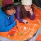 SOL Scout Survival Kit – In Use Inside Tent Two campers using SOL Scout Survival Kit inside a tent with compass and blanket laid out