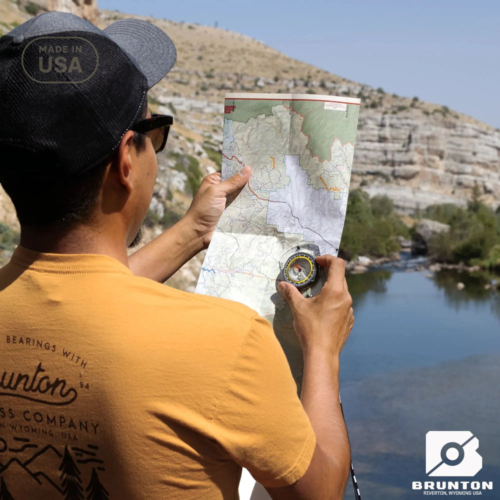 Person using a Brunton compass and topographic map near a river, demonstrating real-world navigation outdoors.