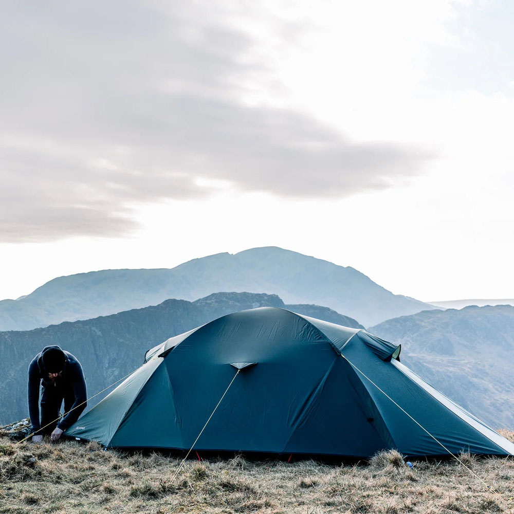 Terra Nova Quasar tent pitched on grassy mountainside during setup, with dramatic hills and cloudy sky in the distance.