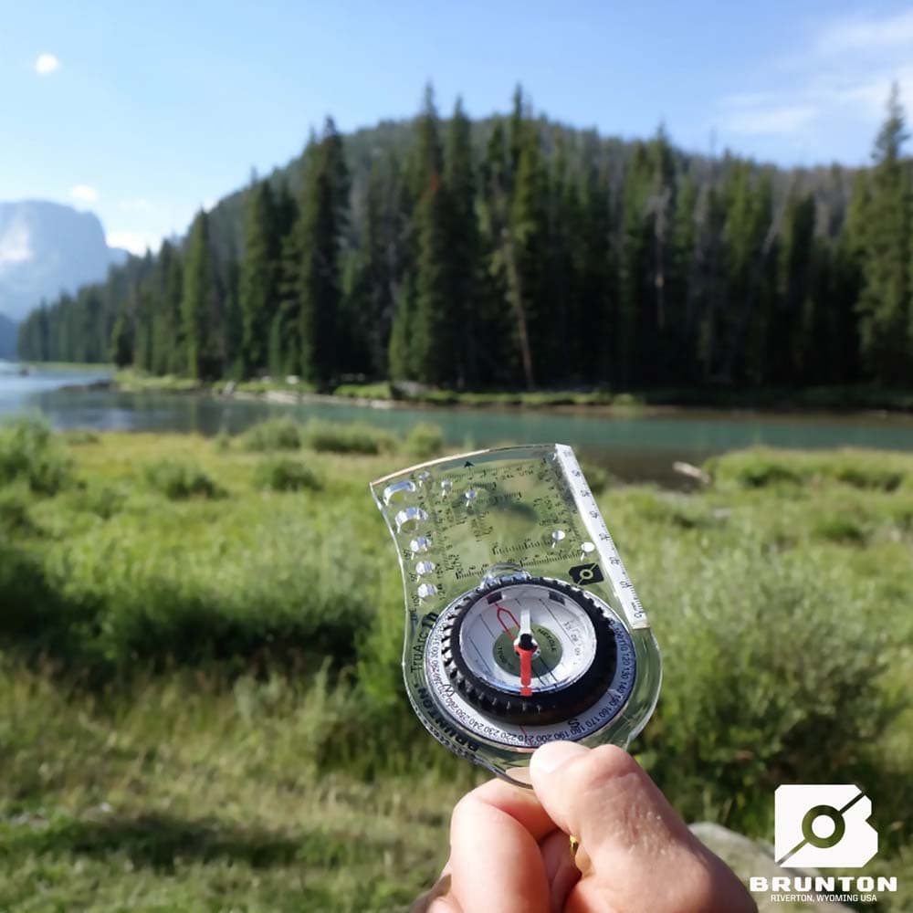 Brunton TruArc 10 Compass in the Wilderness Hand holding the Brunton TruArc 10 compass with a scenic forest and river background