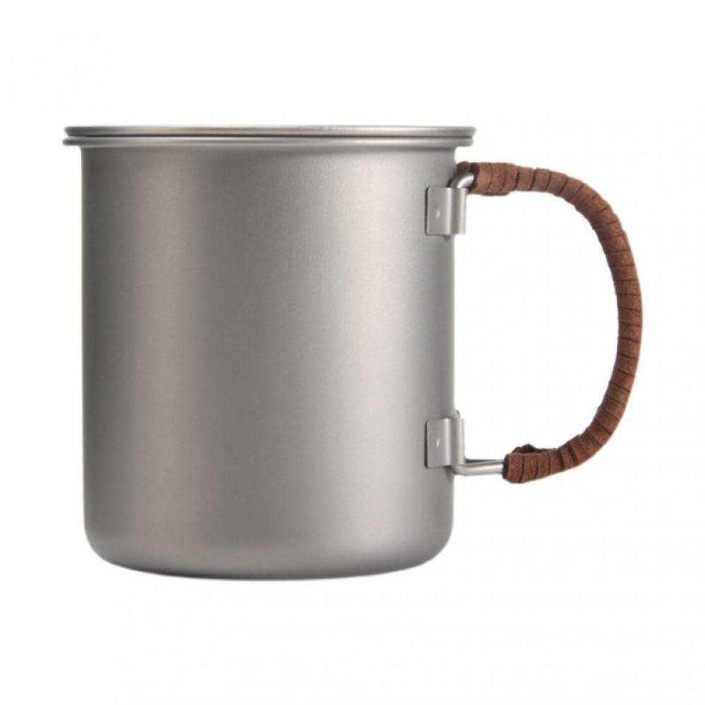 A silver titanium mug with a foldable handle and leather rope attachment, sitting on a rock beside a campfire.