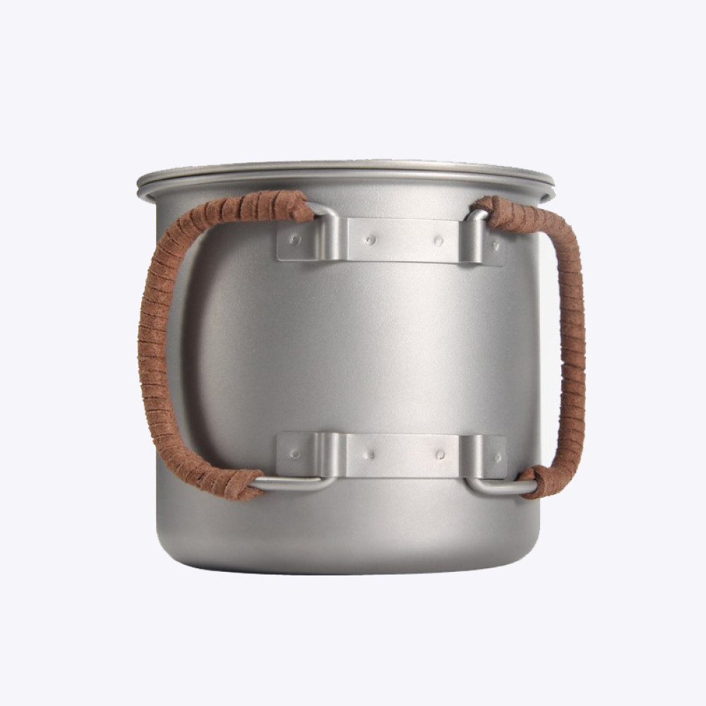 A silver titanium mug with a foldable handle and leather rope attachment, sitting on a rock beside a campfire.